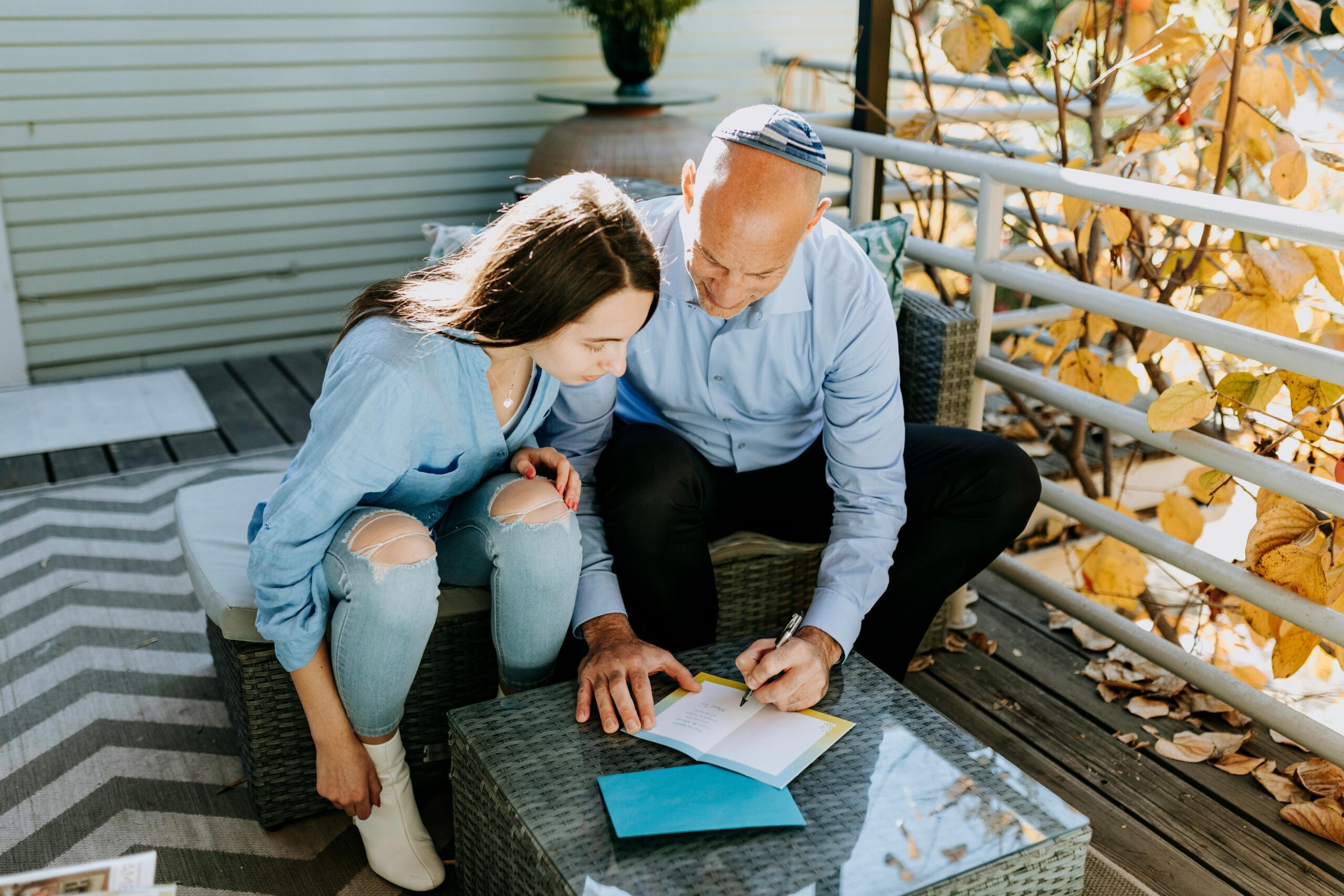 A young woman and an older man, both wearing light blue shirts, sit together on outdoor wicker furniture. The man points at something in an open card while the woman looks on attentively. Yellow leaves are visible nearby.