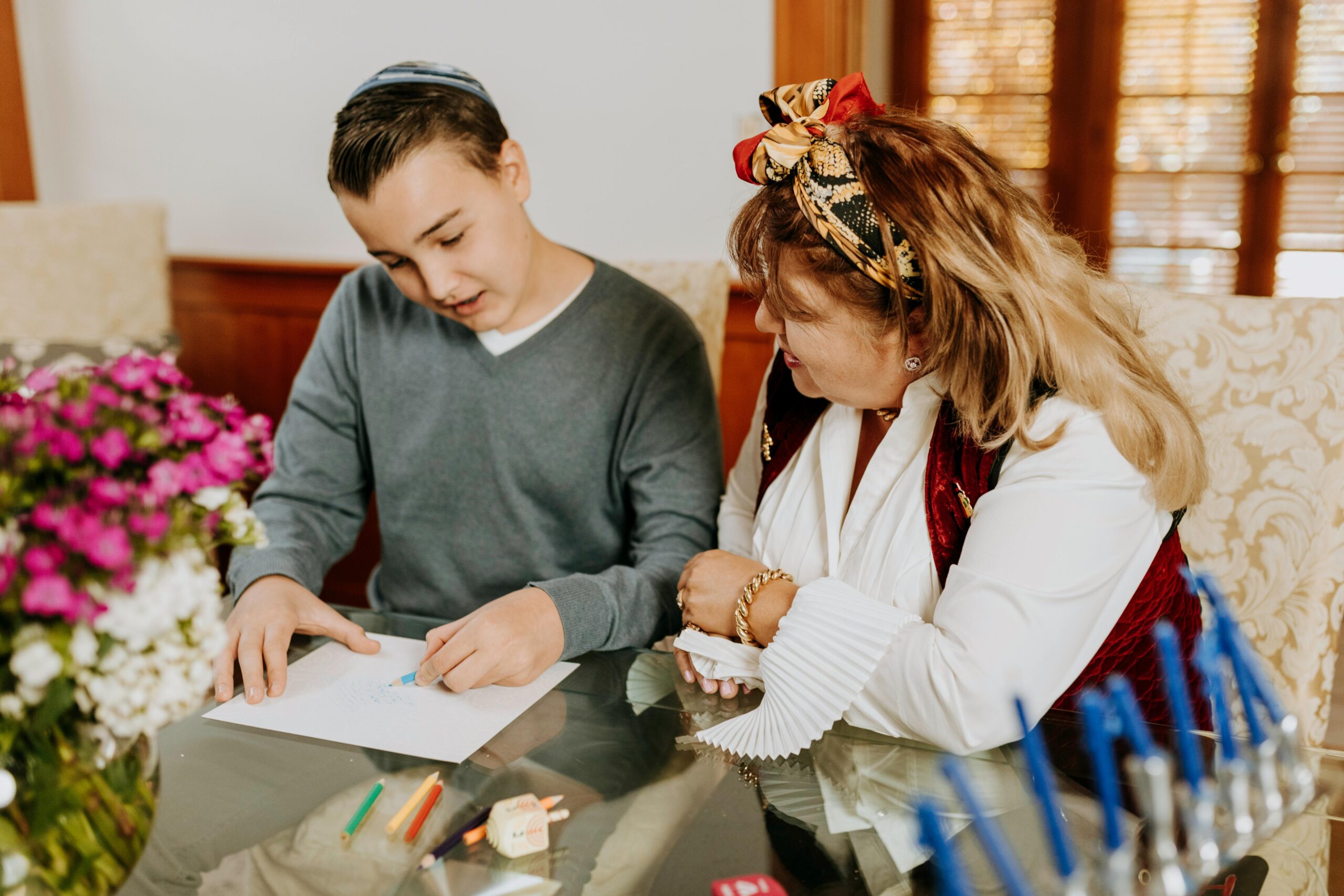 A teenage boy wearing a kippah draws on paper at a table beside a woman in festive attire. Colored pencils, a dreidel, and a menorah with unlit candles are on the table. Flowers are in the foreground.