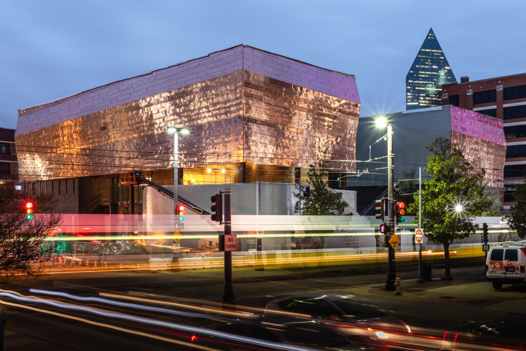 Evening view of the Dallas Holocaust and Human Rights Museum with its copper-colored exterior, city lights, and blurred motion of traffic in the foreground.