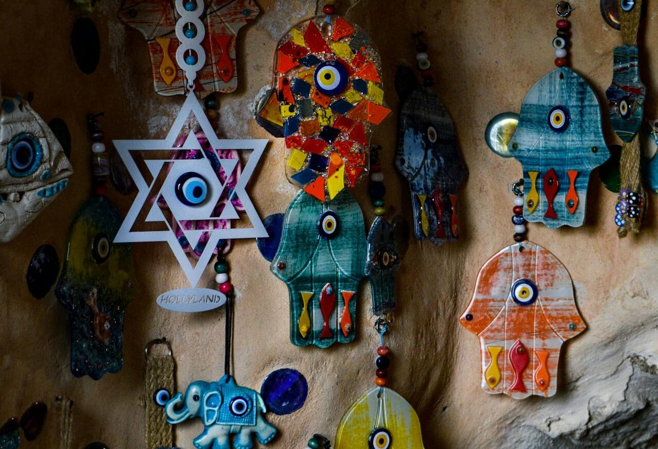 Colorful hanging amulets including Hamsa hands, a Star of David with evil eye, and decorative charms displayed on a wall.