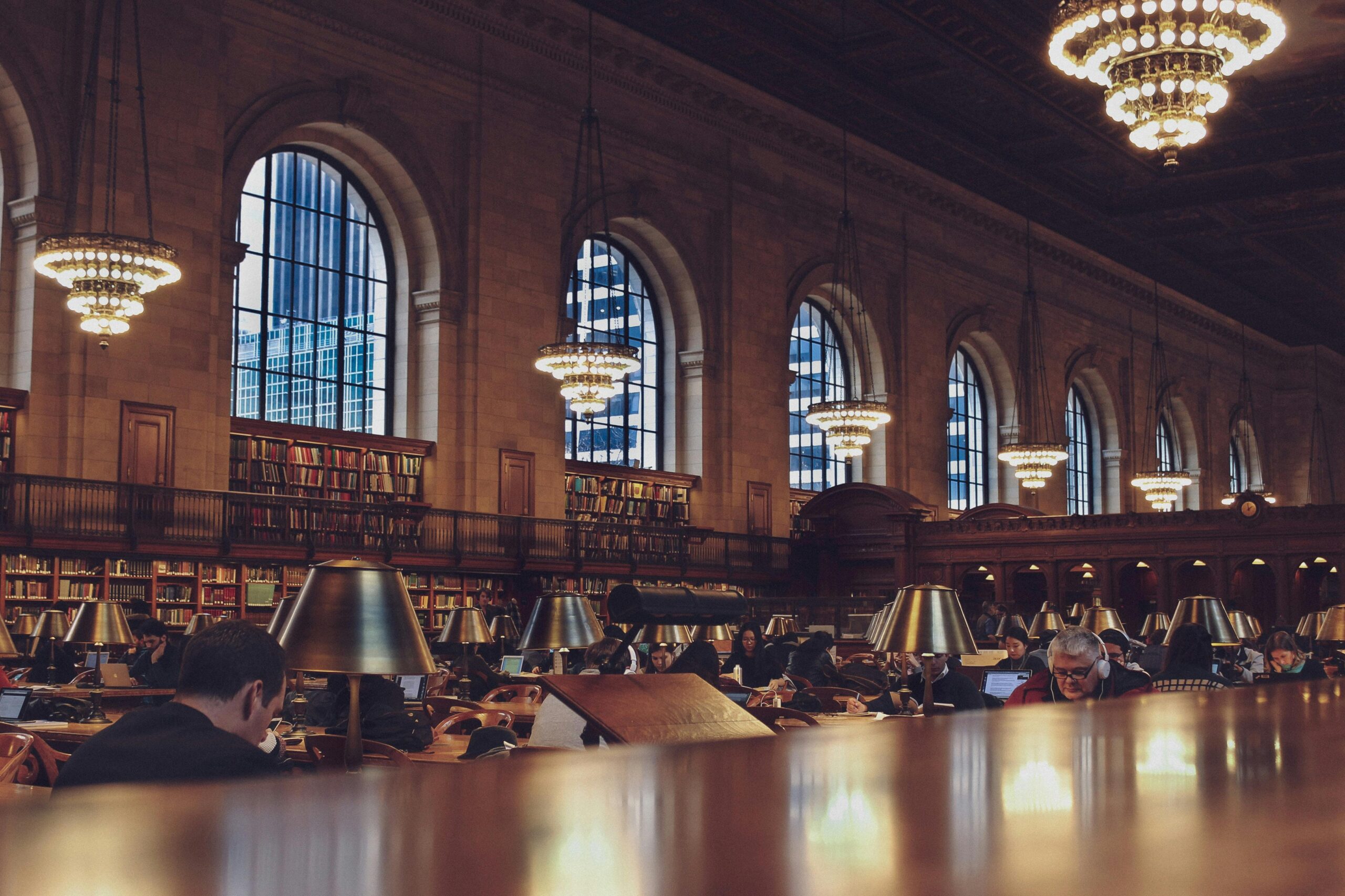 Interior of a grand library reading room with tall arched windows, chandeliers, bookshelves, and people studying at long wooden tables with lamps.