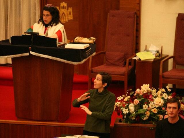 Interpreter using sign language during a synagogue service, with a rabbi at the bimah and congregants seated nearby.